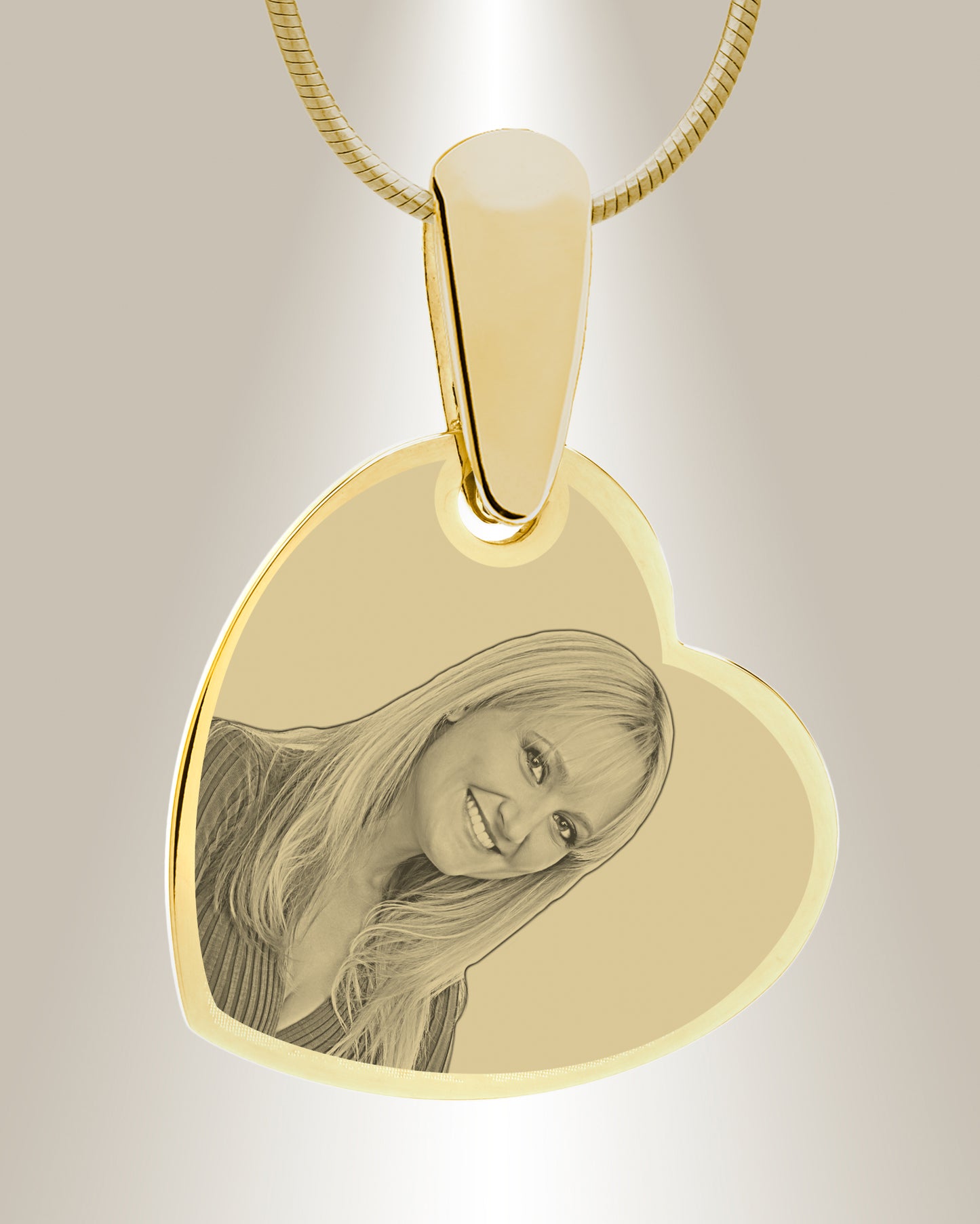A gold-colored heart-shaped photo pendant with an engraved image of a smiling person. The pendant is attached to a gold-colored chain.