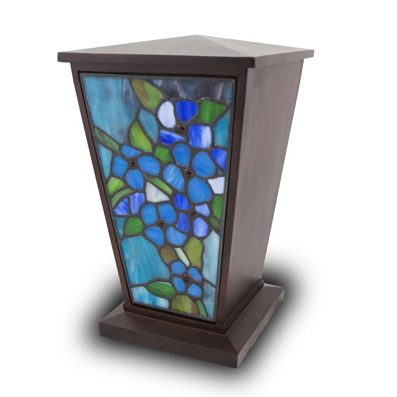 A tall, dark wood cremation urn with a tapered tower shape, featuring a beautiful stained glass front panel of blue and green forget-me-not flowers.