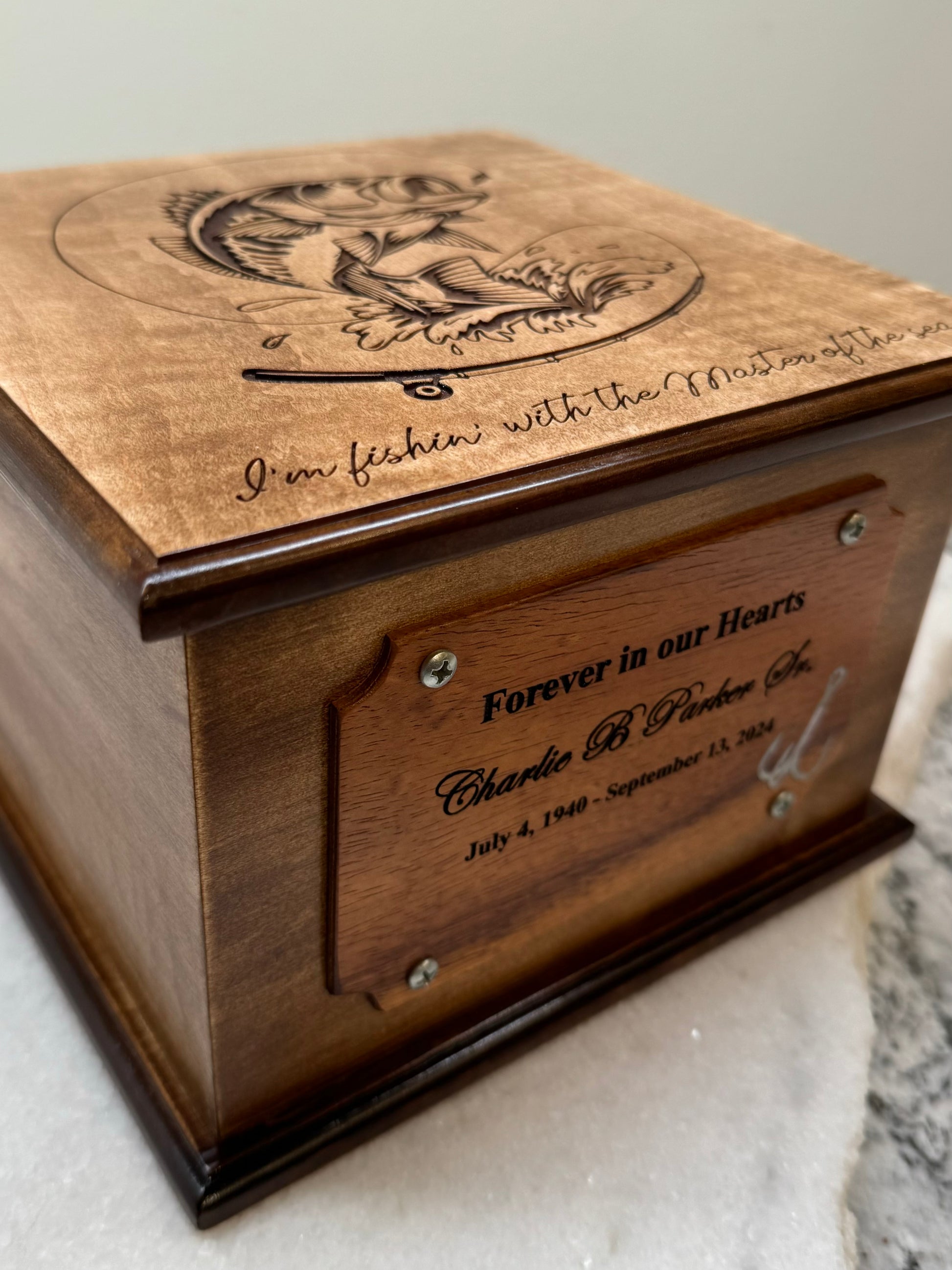 A wooden urn with engraved bass fish on top and a boat on the side, accompanied by the text 'mother of pearl fishhook' on the nameplate.