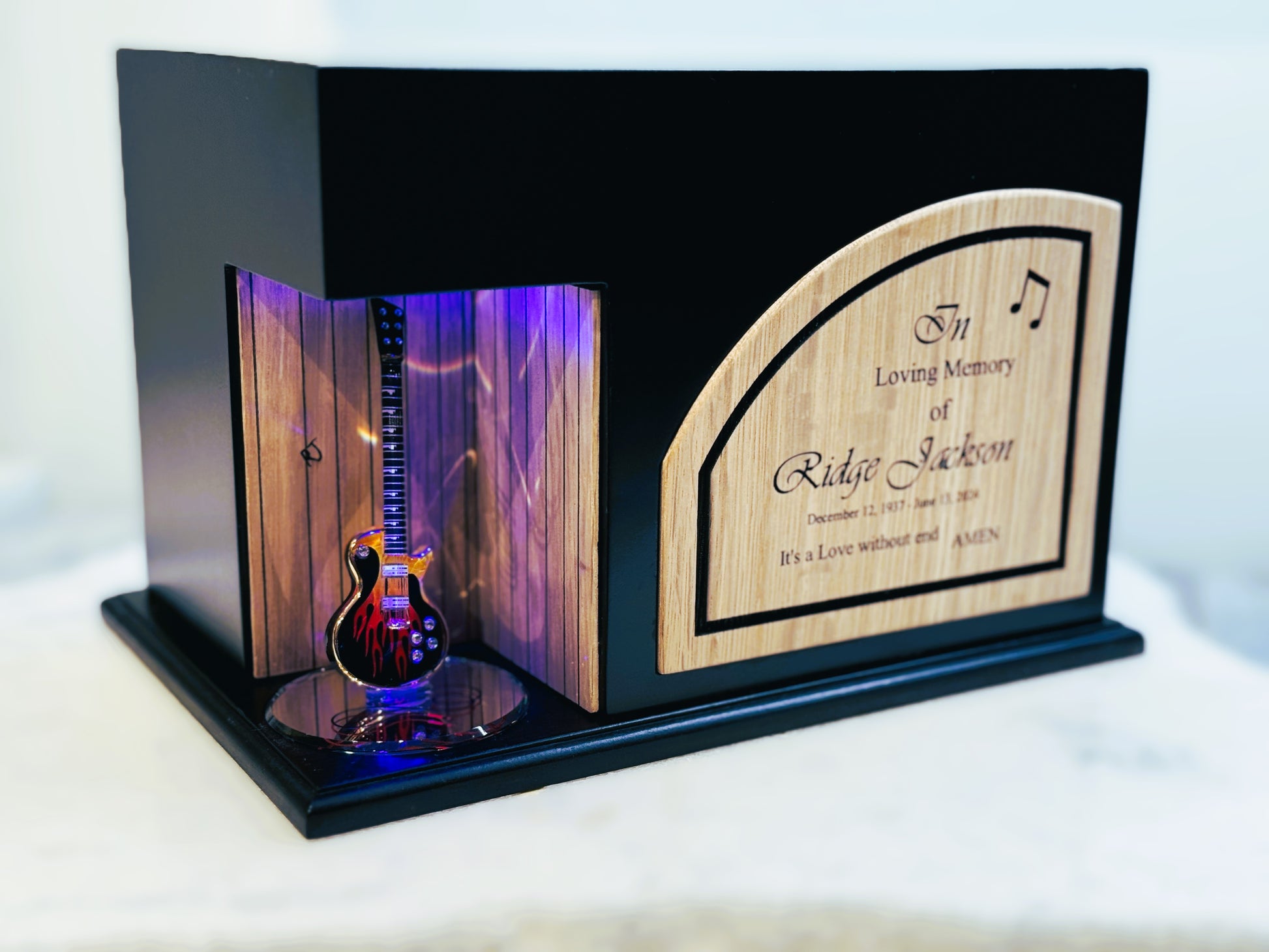 A guitar-shaped urn with a lighted section showcasing the instrument, set within a wooden frame with a name plate.