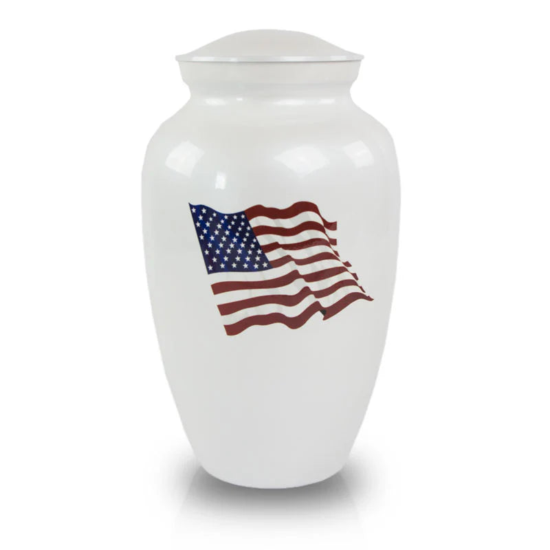 A white brass cremation urn with a engraving of the American flag waving in the wind.