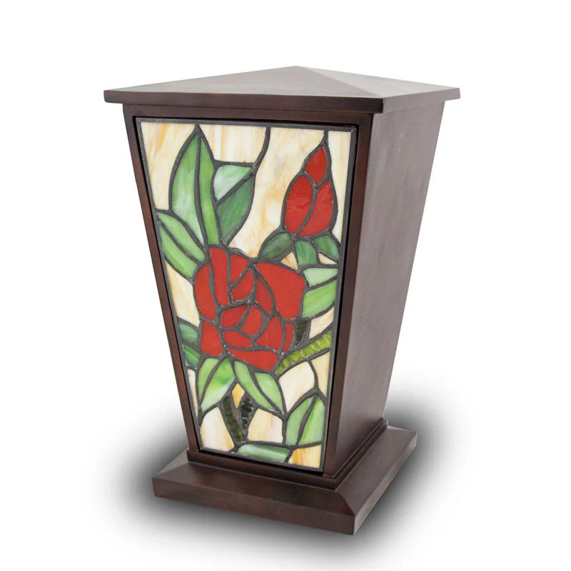 A tall dark wood cremation urn with a tapered tower shape, featuring a stained glass front panel of a red rose and green leaves.