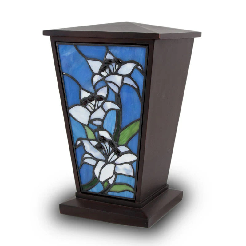 A tall, dark wood cremation urn with a tapered tower shape, featuring a beautiful stained glass front panel of blue and white lily flowers.