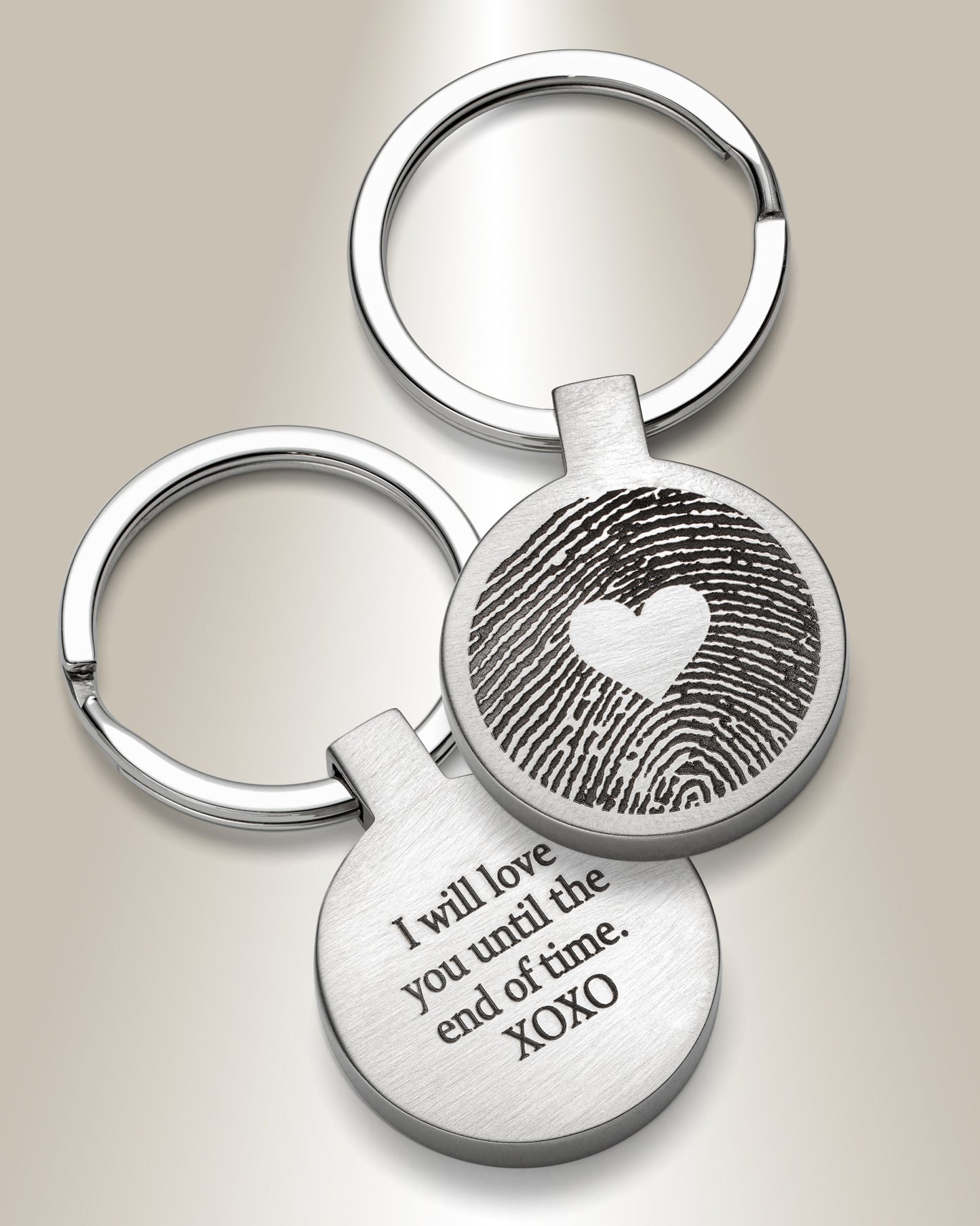 Two stainless steel keychains, one with a fingerprint engraved in the center and a heart shape above it, and the other with text that reads 'I will love you till the end of time. XOXO'.