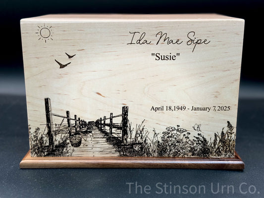 A handcrafted wooden urn with an engraved image of a dock and reeds, and the text 'Ida Mae Spie "Susie"', along with dates 'April 18, 1949 - January 7, 2025'. The urn has a sun illustration at the top and appears to have a walnut bottom.