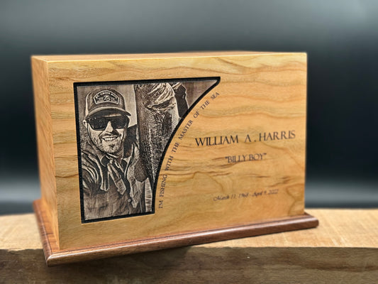 A wooden photo urn with an engraved image of a person wearing a hat and sunglasses on the front, with a walnut bottom and maple inlaid centerpiece.