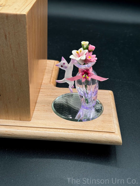 A decorative pet urn featuring a cat figurine with a vase and engraved dandelions, set on a wooden platform.