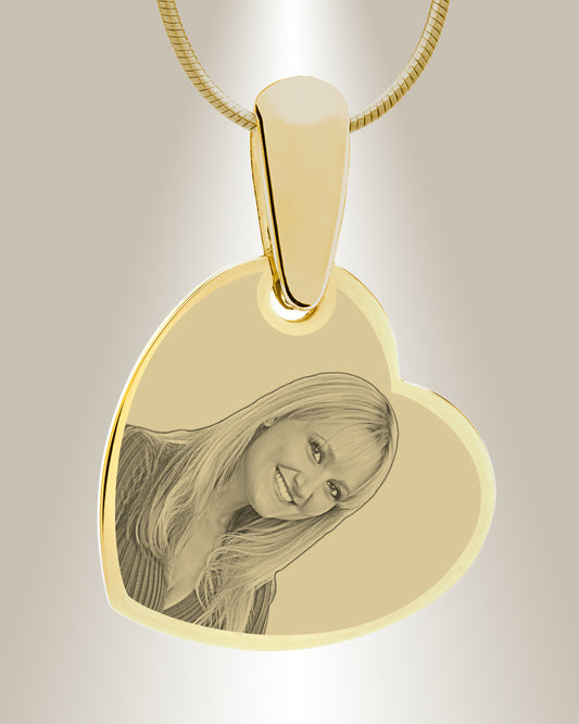 A gold-colored heart-shaped photo pendant with an engraved image of a smiling person. The pendant is attached to a gold-colored chain.