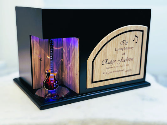 A guitar-shaped urn with a lighted section showcasing the instrument, set within a wooden frame with a name plate.