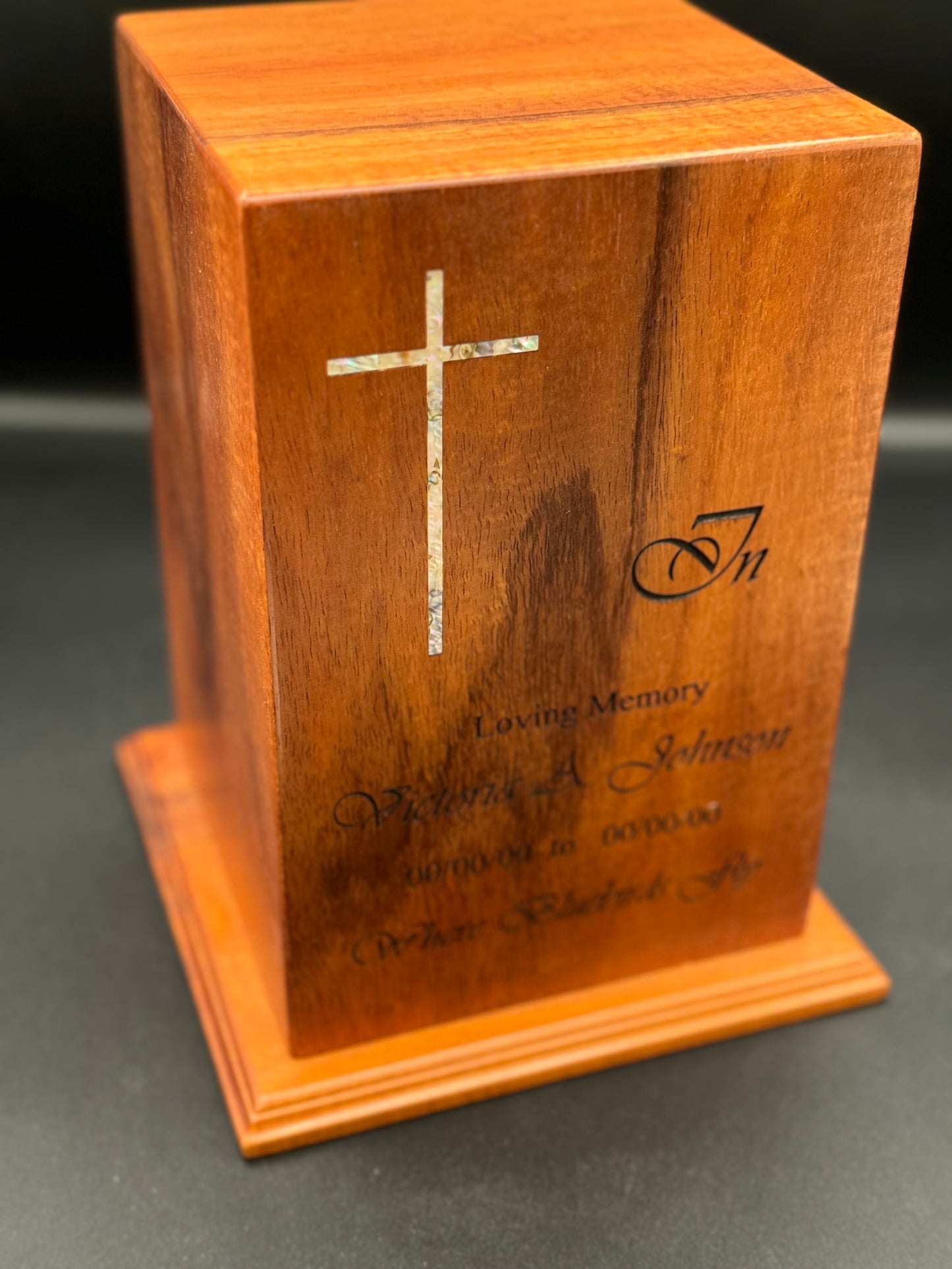A wooden urn with a mother of pearl inlay in the form of a cross, along with engraved text on its surface.