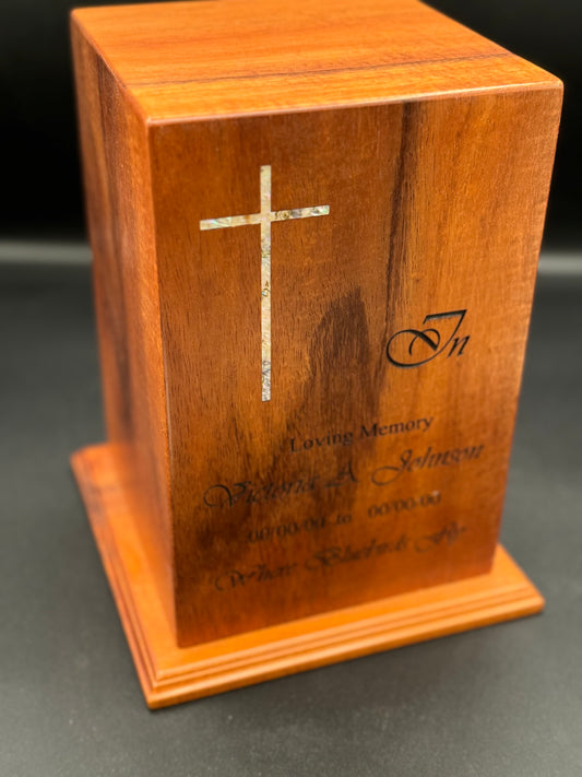 A wooden urn with a mother of pearl inlay in the form of a cross, along with engraved text on its surface.