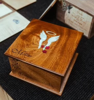 A wooden urn with a mahogany finish, featuring an inlay of mother of pearl depicting angel wings and a halo, surrounded by abalone shell and red stone designs.