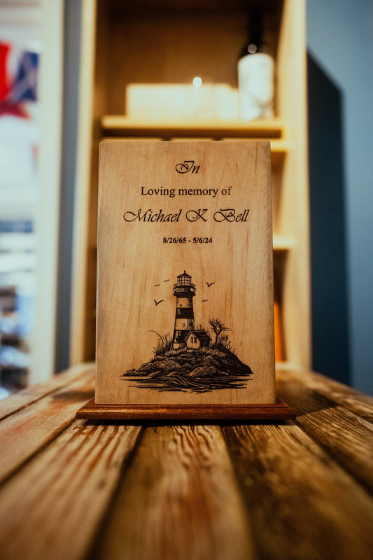 A wooden cremation urn with a laser-engraved lighthouse, surrounded by a detailed background scene of rocks and seagulls.