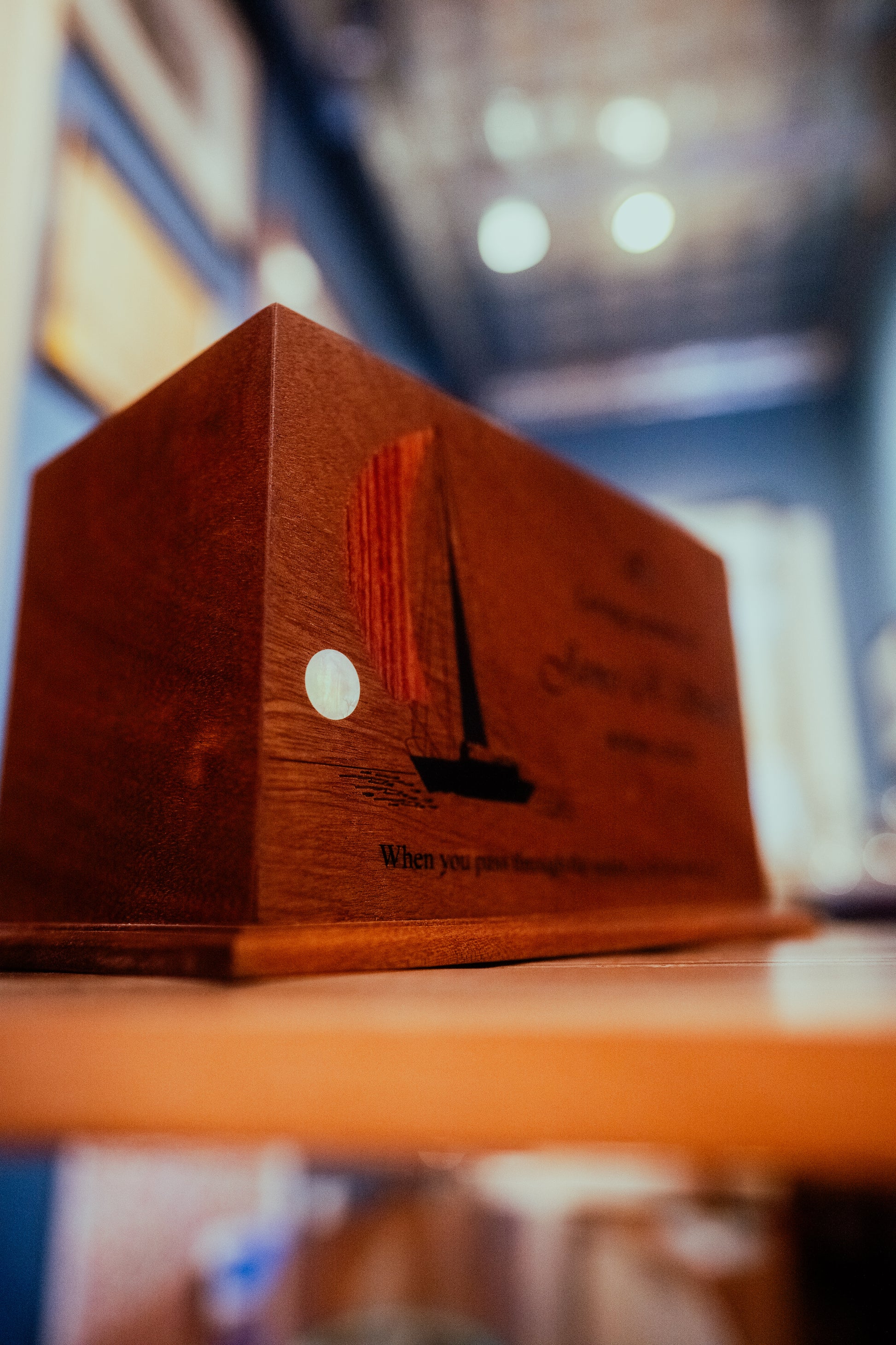 A wooden memorial urn with an elegant sailboat silhouette design, featuring a sunset in the background, displayed on a shelf.
