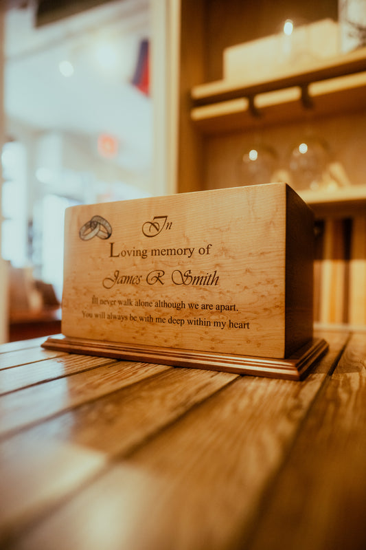 A wooden memorial urn with engraved text, placed on a wooden surface, with a blurred background.