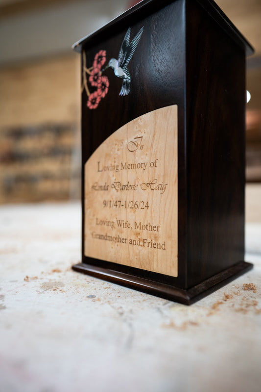 A wooden urn with a curved nameplate made of maple wood, featuring an inlay of a hummingbird and butterfly on the front, set against an indoor background.